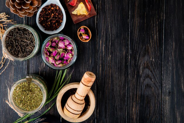 Top view of various spices and herbs dry black tea leaves, peppermint, rose buds, clove spice and black peppercorns in glass jars on black wood with copy space