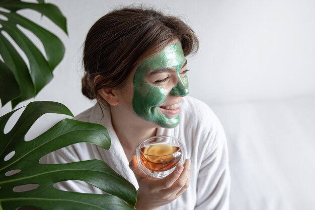 Attractive young woman with a green cosmetic mask on her face and in a white robe with tea in her hands, the concept of spa procedures.